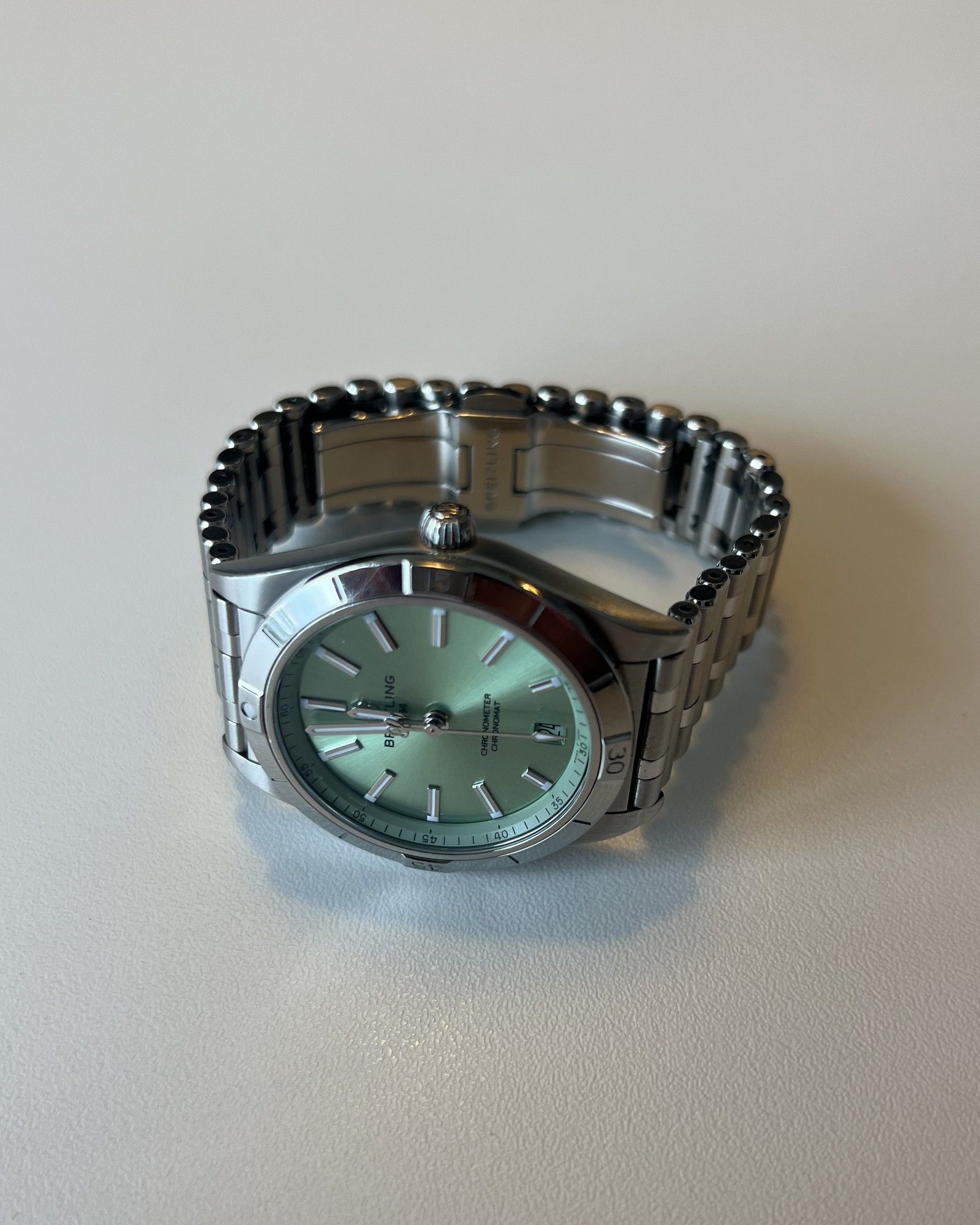 Phone photo of a Breitling wristwatch with a mint green dial and stainless steel bracelet on a light grey table