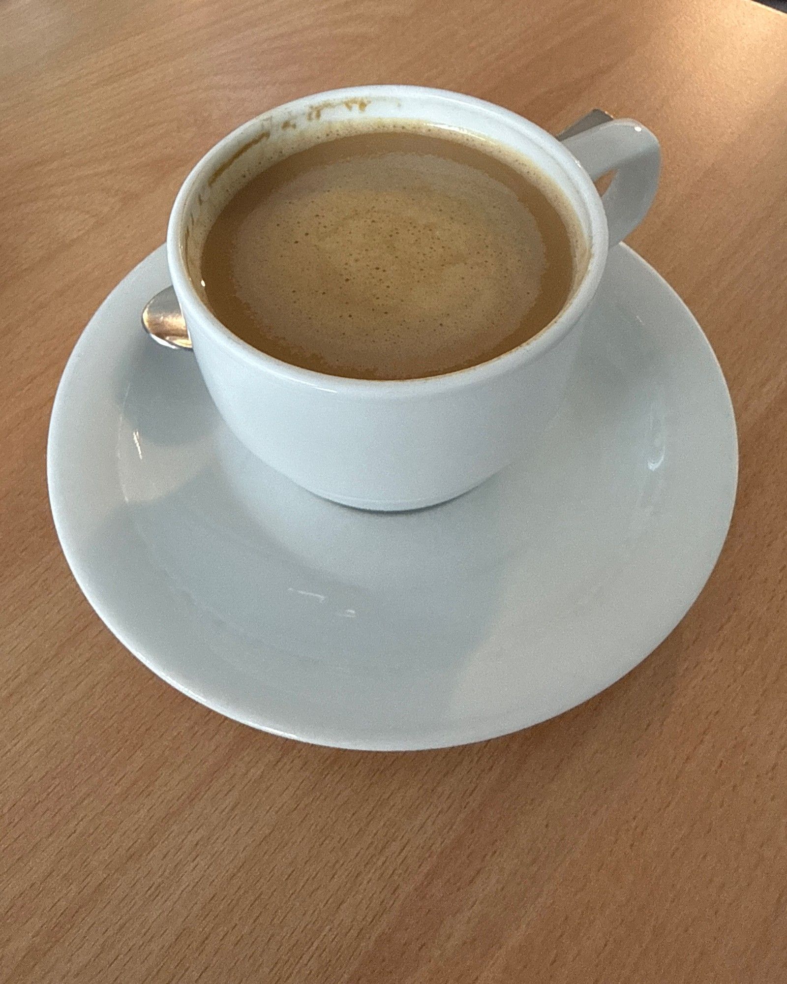 Phone photo of a white coffee cup with saucer on a wooden table, filled with milky coffee