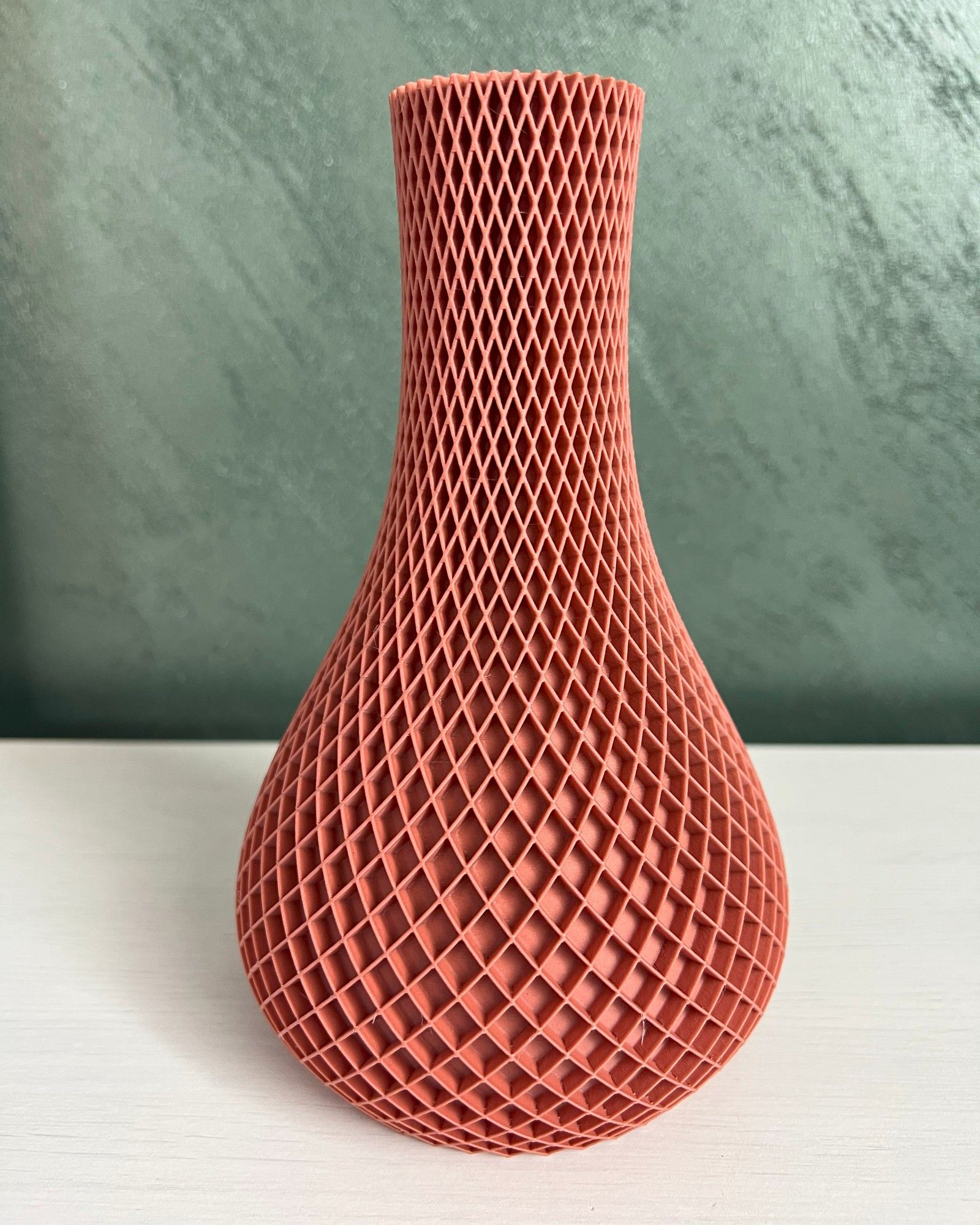 Handyfoto einer terrakottafarbenen, 3D-gedruckten Vase mit Rautengitter auf einem weißen Sideboard vor einer grünen Wand